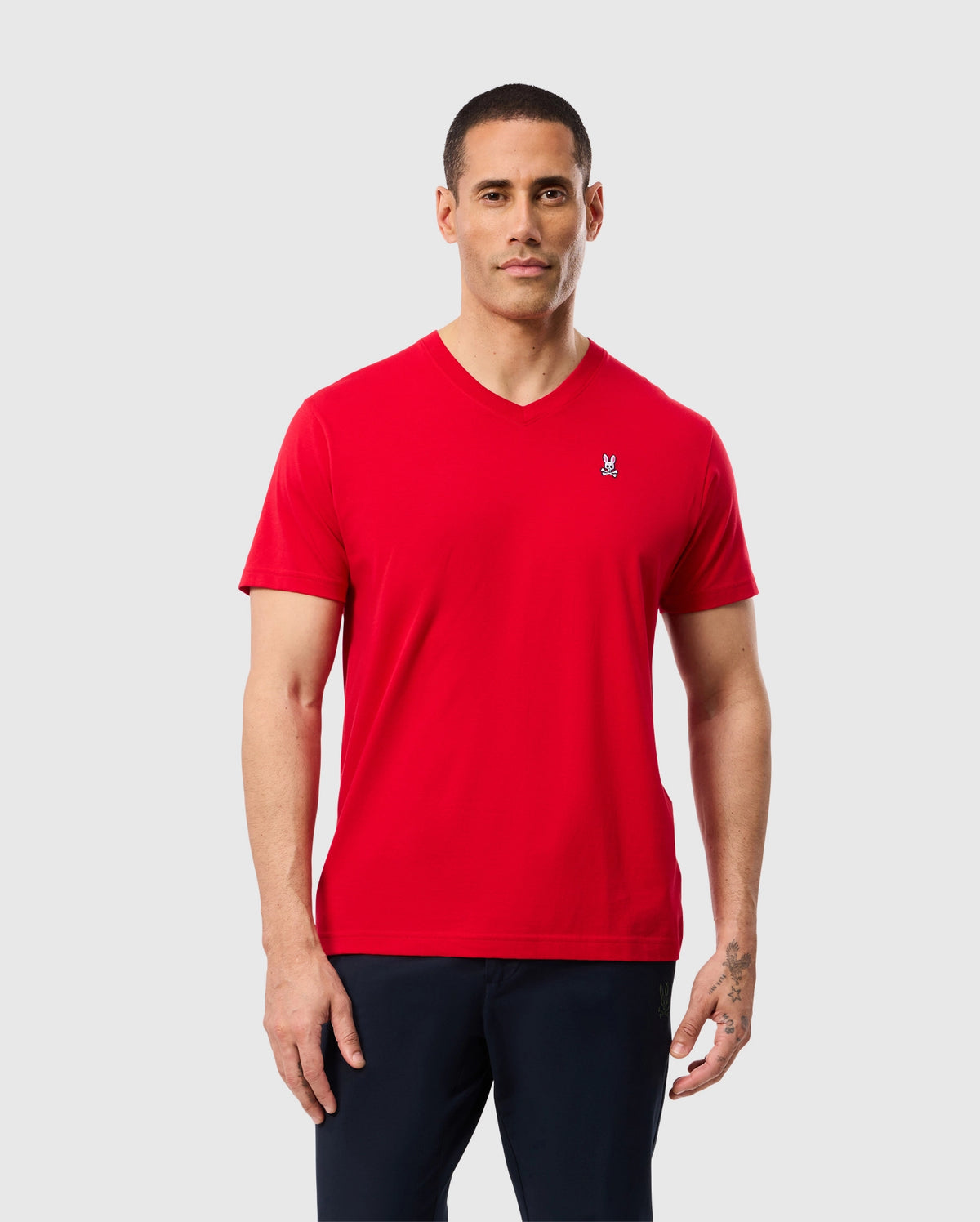 A man with short hair is wearing a bright red Psycho Bunny MENS CLASSIC V NECK TEE - B6U100CRPC, made from 100% Pima cotton, featuring a small white embroidered Psycho Bunny logo on the left chest. He is also wearing dark pants and stands against a plain light gray background.