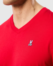 A close-up of a person wearing the Psycho Bunny MENS CLASSIC V NECK TEE - B6U100CRPC, made from 100% Pima cotton in red. The left chest area features an embroidered Psycho Bunny logo with a white cartoon bunny sporting black eye patches and crossbones beneath it. The background is plain and light grey.