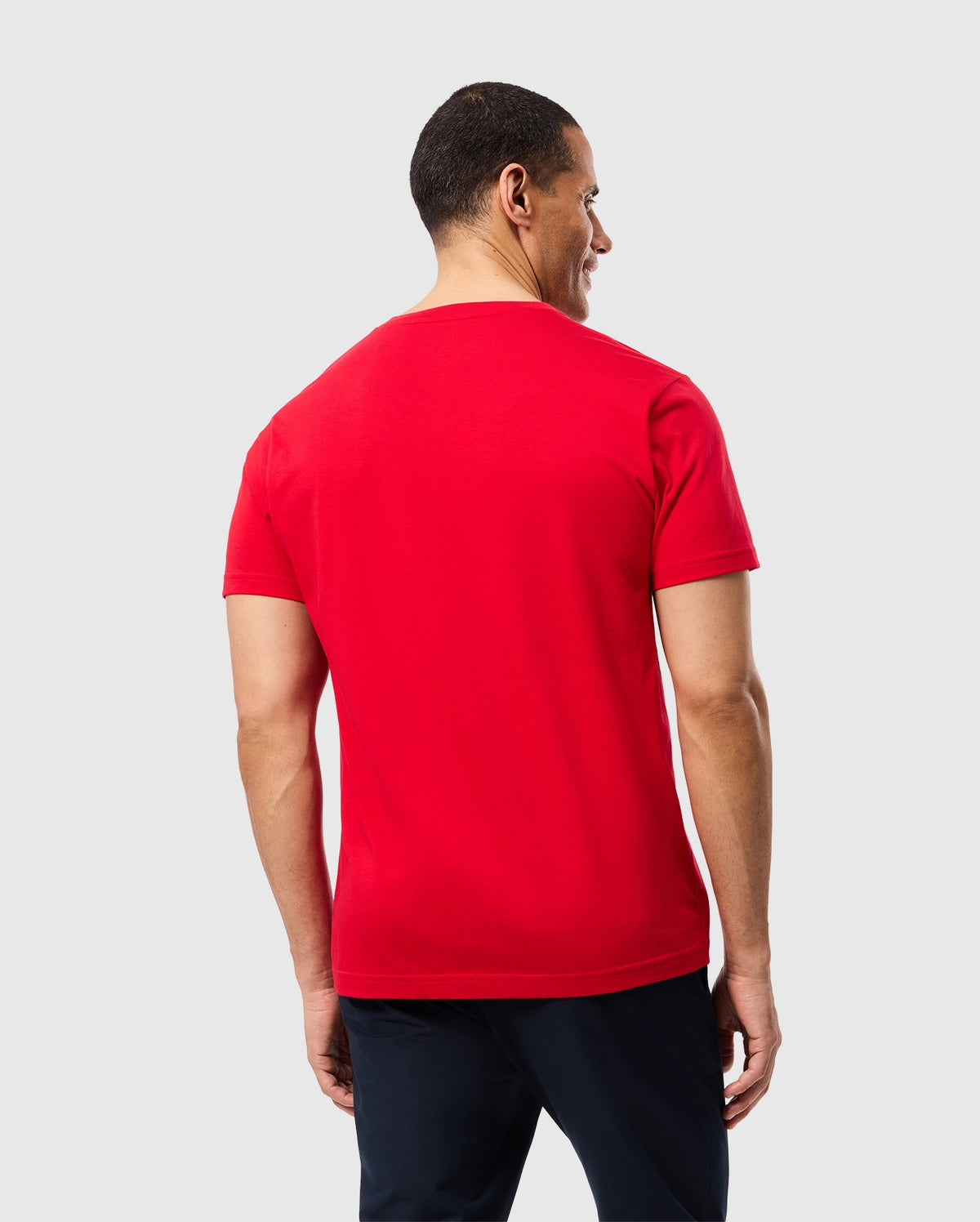 A man with short black hair is seen from the back, wearing the MENS CLASSIC V NECK TEE - B6U100CRPC in bright red by Psycho Bunny, paired with dark pants. He stands against a plain light gray background, slightly turned to his left, with his right arm hanging down and his left hand partially tucked into his pocket.