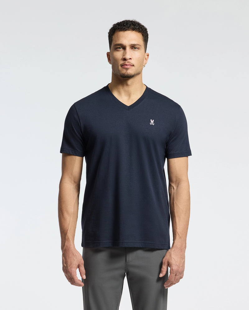 Against a plain backdrop, a person with short curly hair and a neutral expression faces the camera. They're wearing Psycho Bunny's MENS CLASSIC V NECK TEE in dark Pima cotton, featuring a small white logo on the left chest, paired with gray pants.