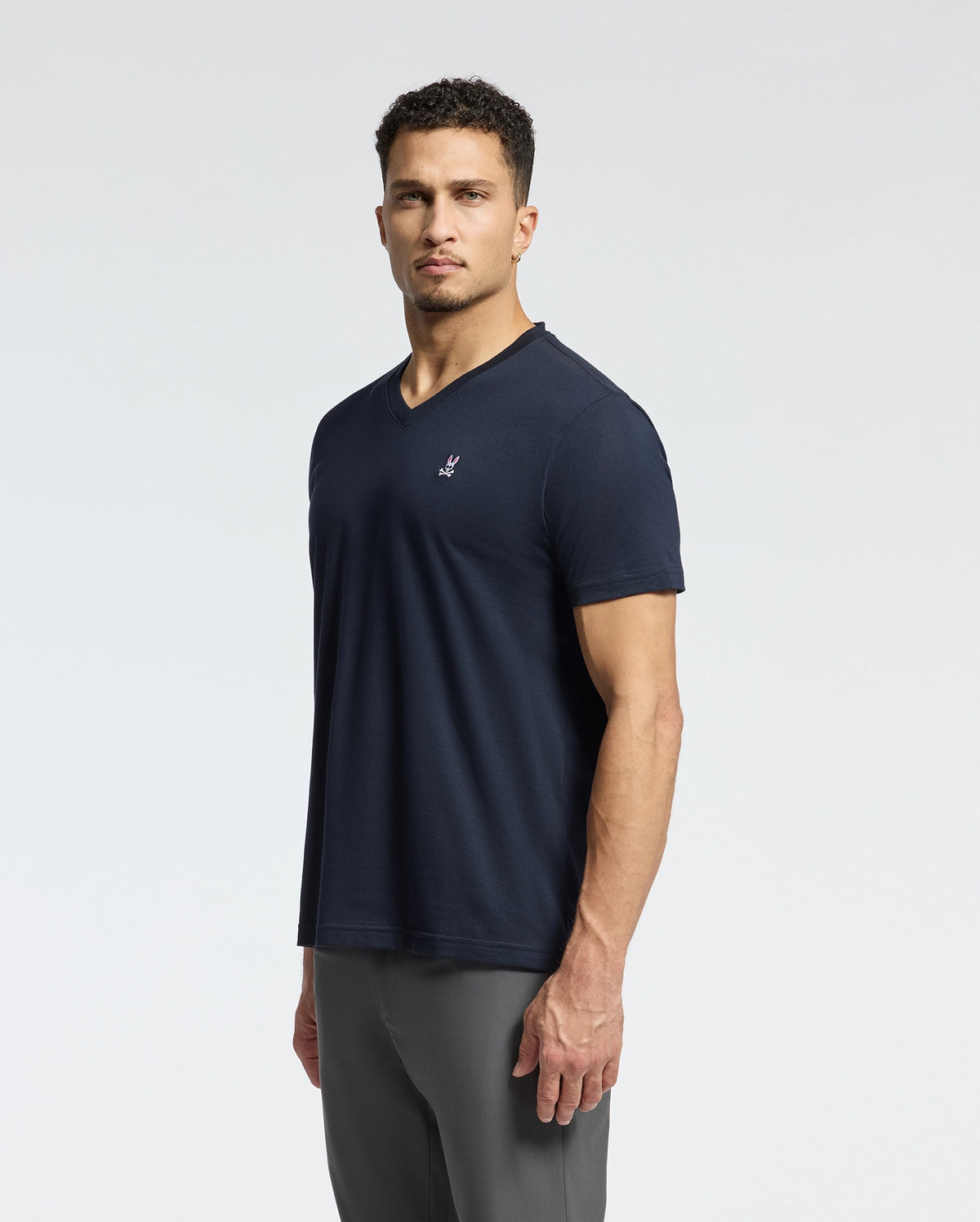 A person with short curly hair is wearing a dark blue Psycho Bunny MENS CLASSIC V NECK TEE - B6U100ARPC made from Pima cotton and gray pants, standing against a plain, light-colored background.