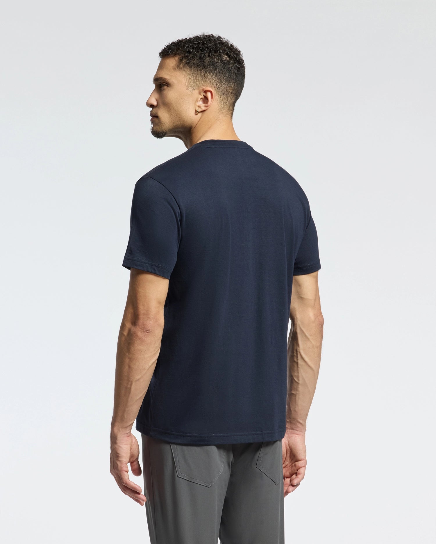 The man with short curly hair stands facing away, wearing a Psycho Bunny MENS CLASSIC V NECK TEE - B6U100ARPC in navy blue and gray pants against a plain light background.
