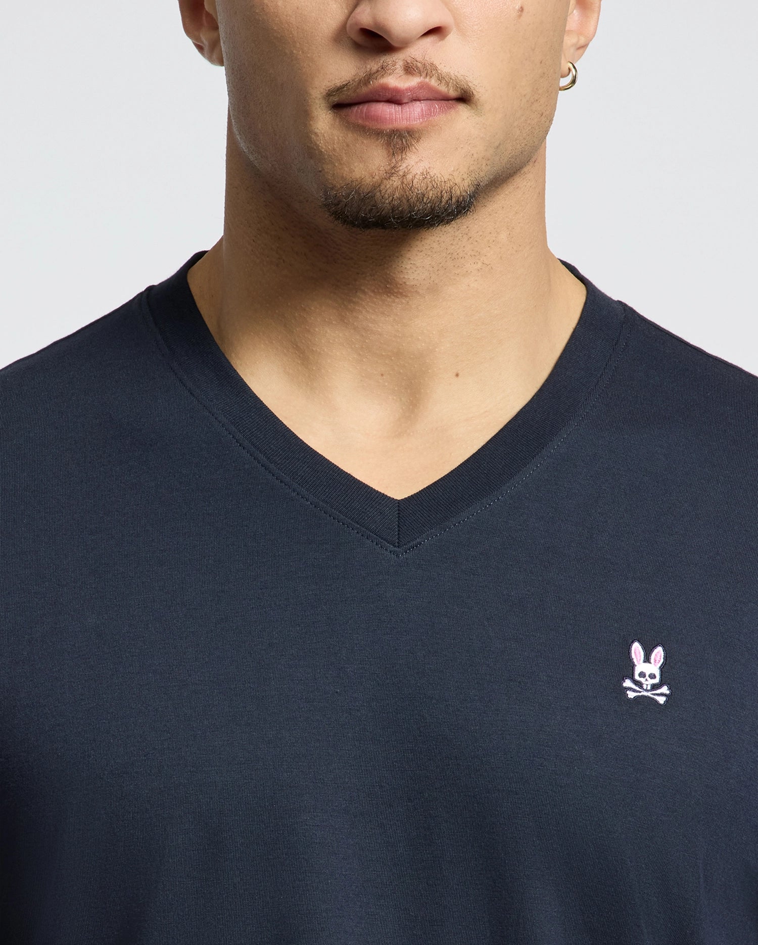 A person is wearing the MENS CLASSIC V NECK TEE by Psycho Bunny, made from soft Pima cotton. It features the iconic pink bunny with crossbones logo on the chest, emphasizing the stylish design of the dark shirt and upper torso.