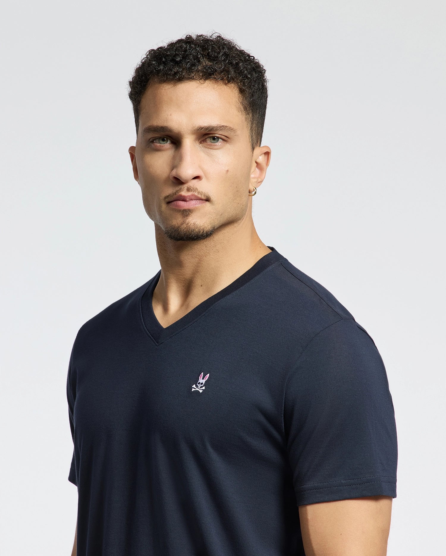 A person with short curly hair is wearing a Psycho Bunny MENS CLASSIC V NECK TEE - B6U100ARPC in navy Pima cotton, featuring an embroidered logo on the left chest, set against a plain white background.