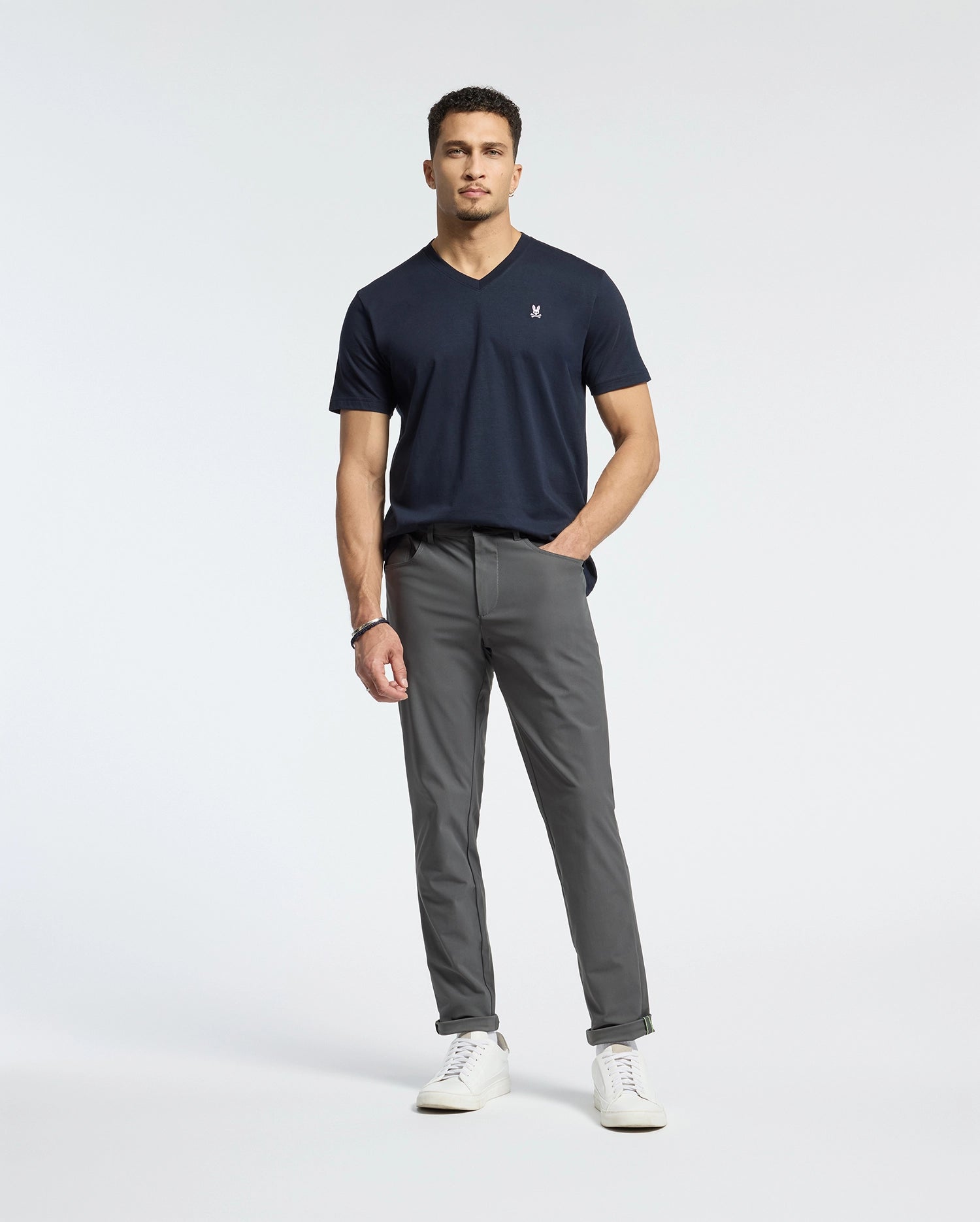 A man stands against a white background wearing gray pants and the Psycho Bunny MENS CLASSIC V NECK TEE - B6U100ARPC in dark navy. Made of Pima cotton, it pairs well with his short curly hair and white sneakers. His right hand is in his pocket, with his left arm by his side.