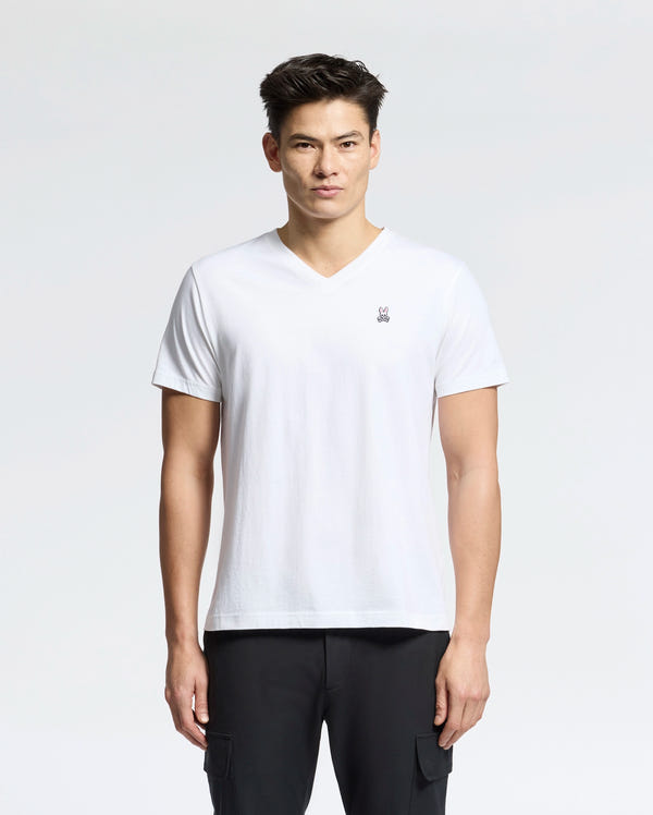 A man with short dark hair is wearing a white MENS CLASSIC V NECK TEE - B6U100ARPC by Psycho Bunny, made in Peru from 100% Pima cotton, featuring a small emblem on the chest. He pairs it with black pants and stands against a light gray background.