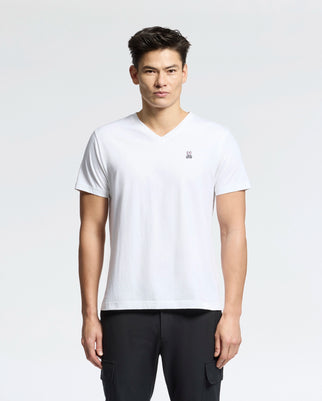 A man with short dark hair is wearing a white MENS CLASSIC V NECK TEE - B6U100ARPC by Psycho Bunny, made in Peru from 100% Pima cotton, featuring a small emblem on the chest. He pairs it with black pants and stands against a light gray background.