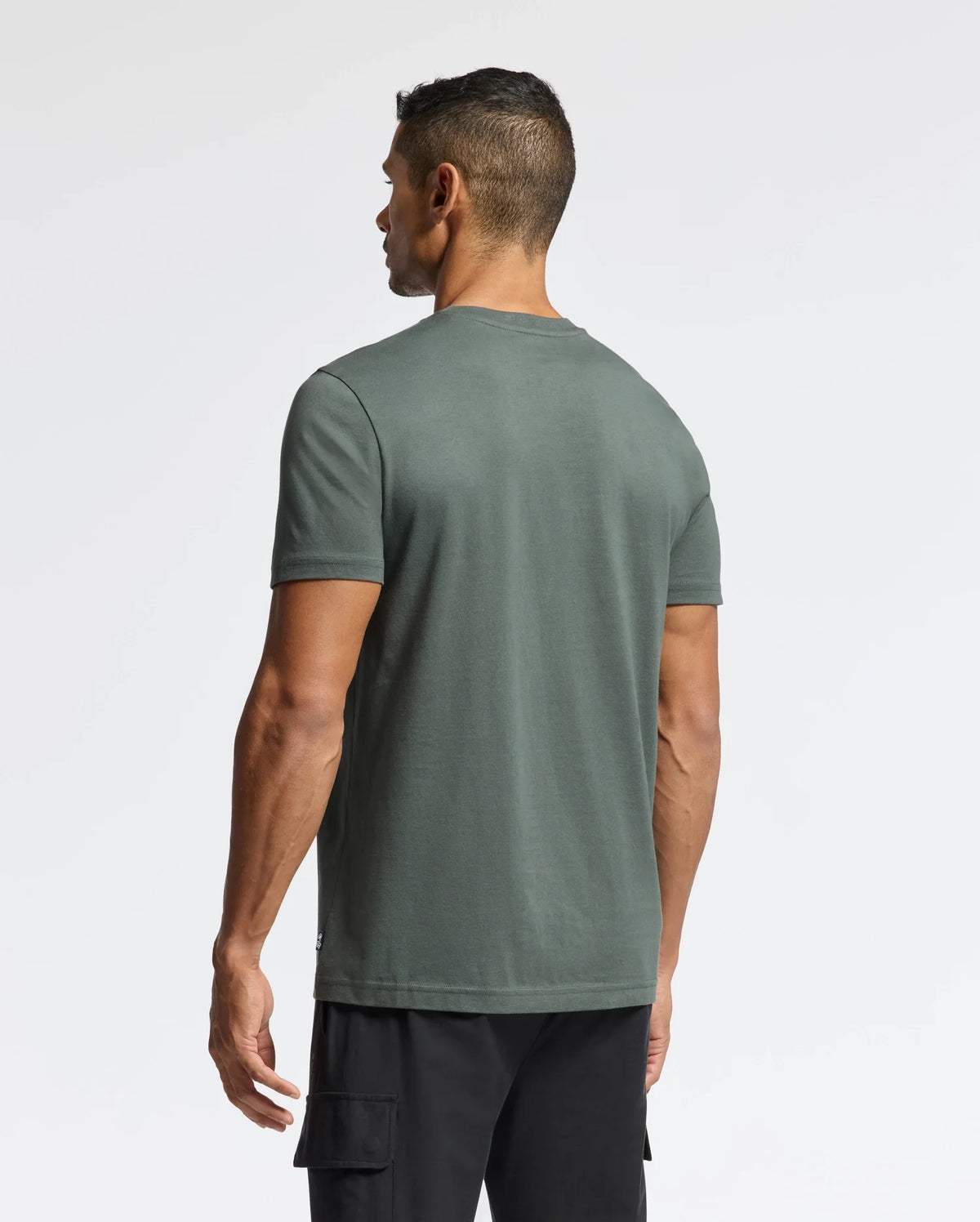 A man is seen from behind, wearing a dark green Psycho Bunny MENS PIERCE EMBROIDERED GRAPHIC TEE - B6U115E200 made of soft Pima cotton paired with black cargo shorts, set against a plain white background.