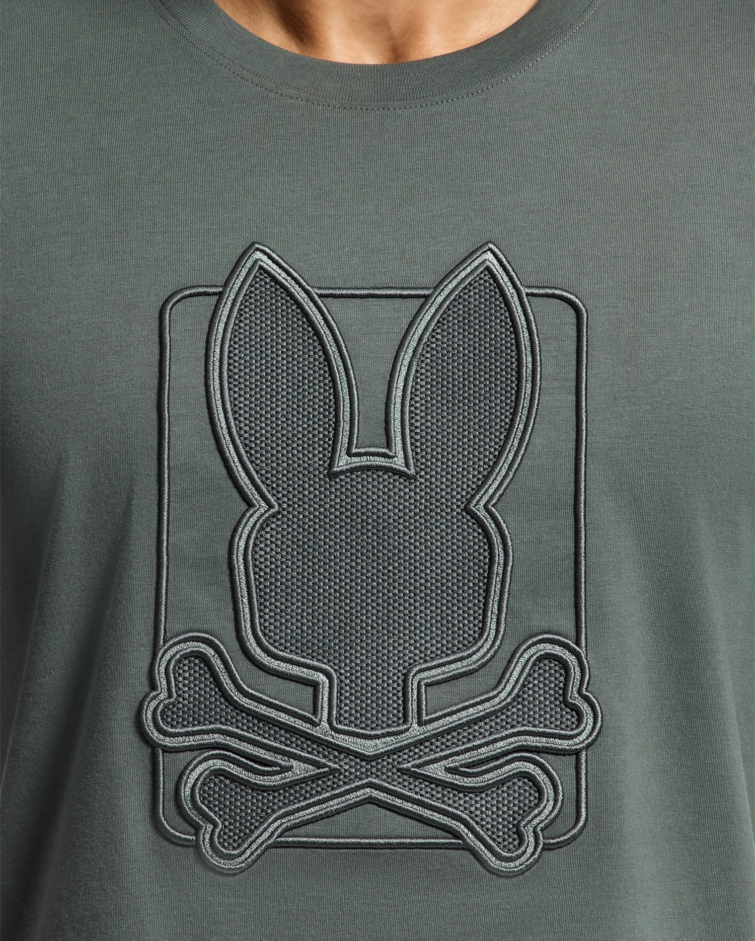 Detailed view of a dark green Psycho Bunny Men's Pierce Embroidered Graphic Tee (B6U115E200) crafted from soft Pima cotton, showcasing an embroidered design of a bunny head with crossed bones beneath. The embroidery features an outline, creating a raised texture on the fabric.