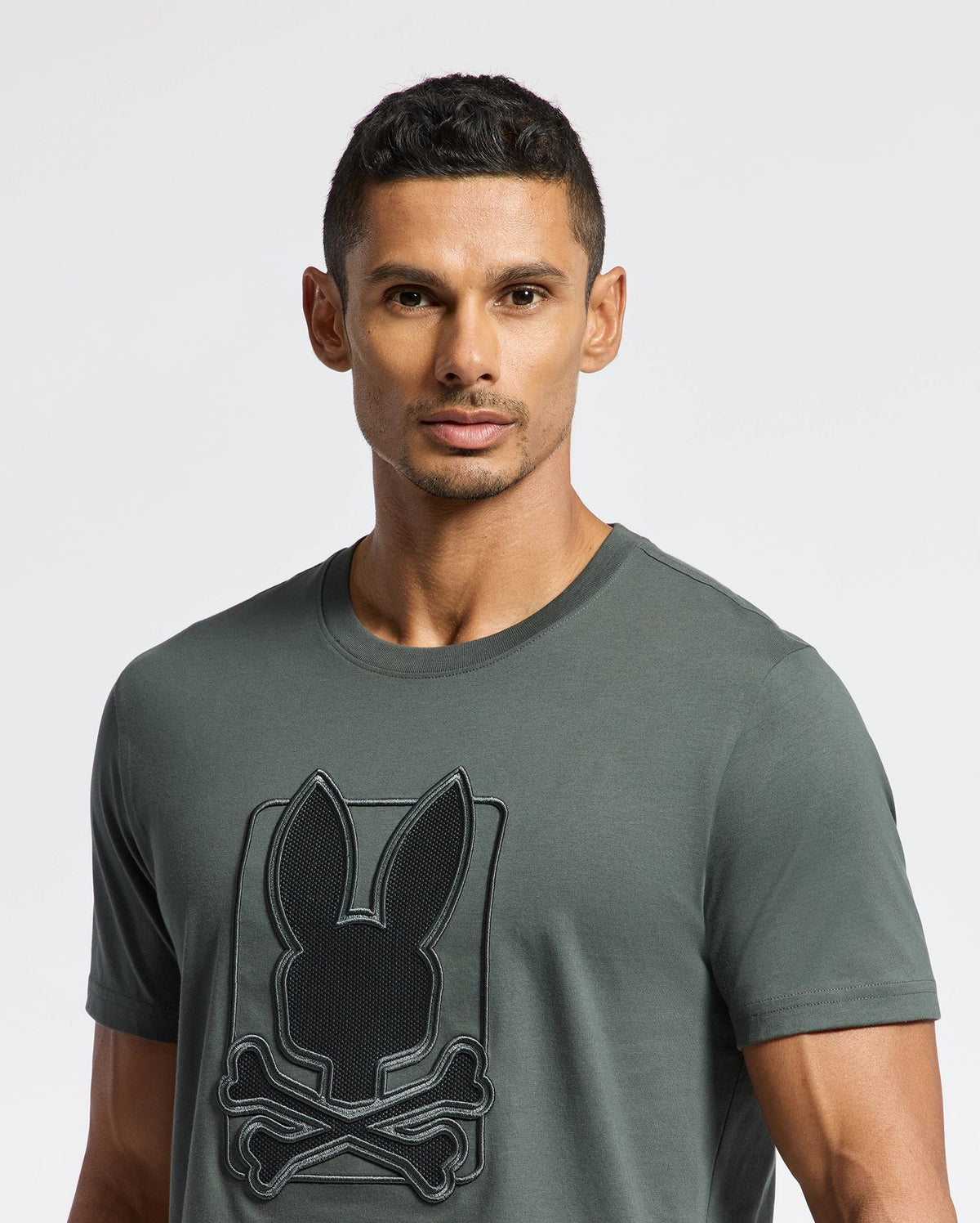 A man with short dark hair is wearing a Psycho Bunny MENS PIERCE EMBROIDERED GRAPHIC TEE - B6U115E200 crafted from soft Pima cotton, which features a stylized design of a bunny and crossed bones. He stands against a plain white background.