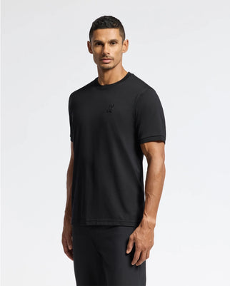 A man in a Psycho Bunny MENS BRANSON FASHION TEE - B6U137E200, crafted from premium Pima cotton, with a small embroidered emblem on the chest, and black pants stands against a light gray background. His hair is short and styled.