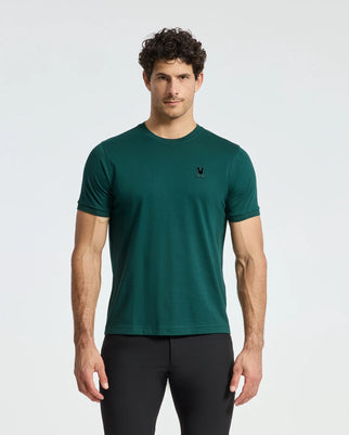 A man with curly hair wears a dark green Psycho Bunny MENS BRANSON FASHION TEE - B6U137E200, featuring a small chest logo, paired with black pants. The background is plain white.