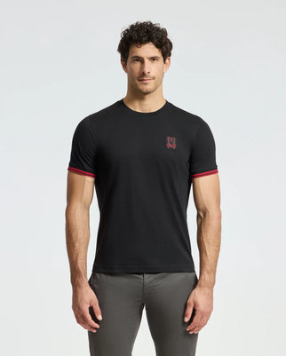 A man wearing a Psycho Bunny MENS PIERCE FASHION TEE - B6U140E200, made from 100% Pima cotton, showcasing a small red logo on the chest and featuring red accents on the sleeves, is paired with gray pants against a plain light background.
