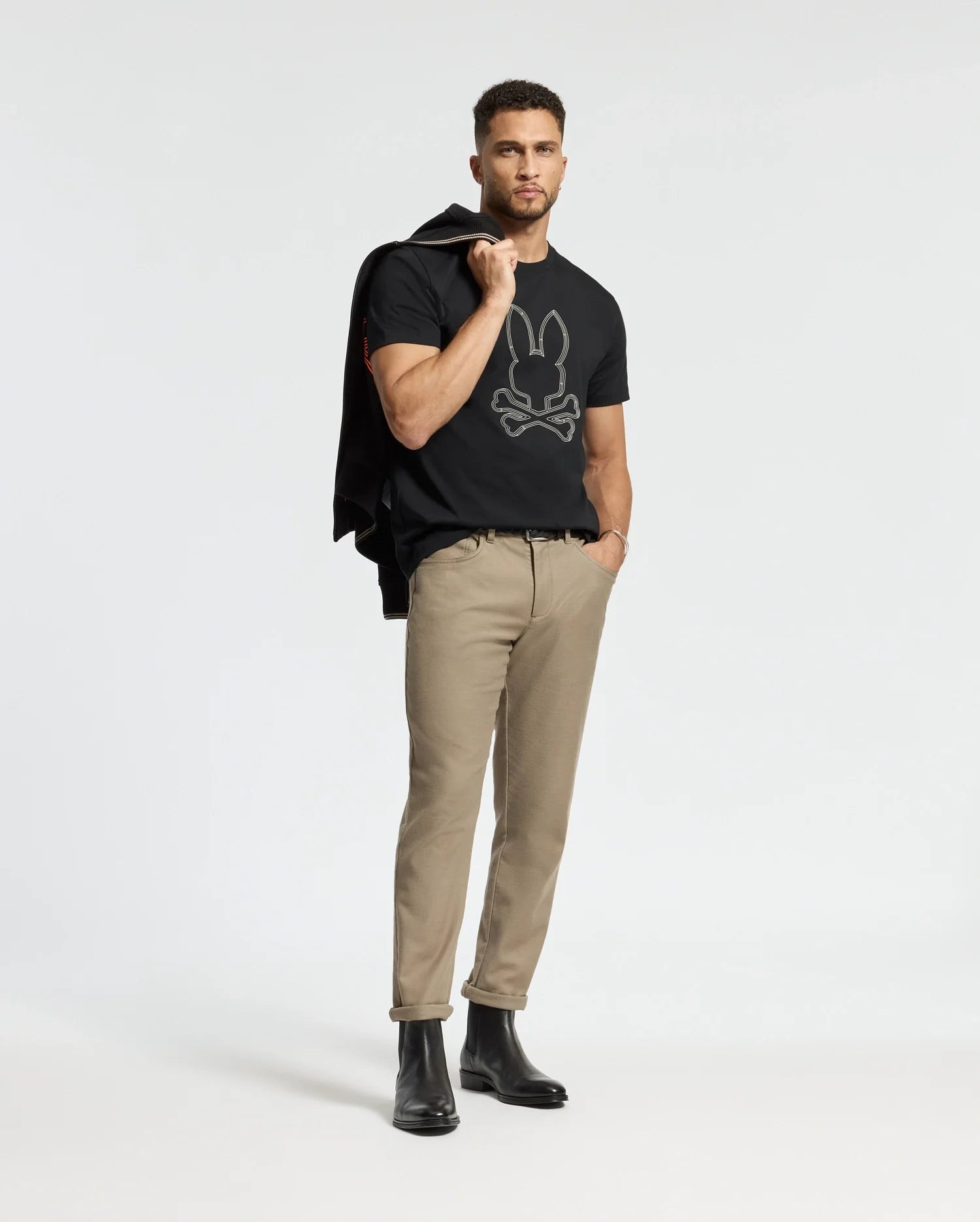A man stands against a plain background, wearing a black Psycho Bunny Tee (B6U211E200), beige pants, and black boots, with a Monarc jacket slung over his shoulder, creating a relaxed yet stylish look.