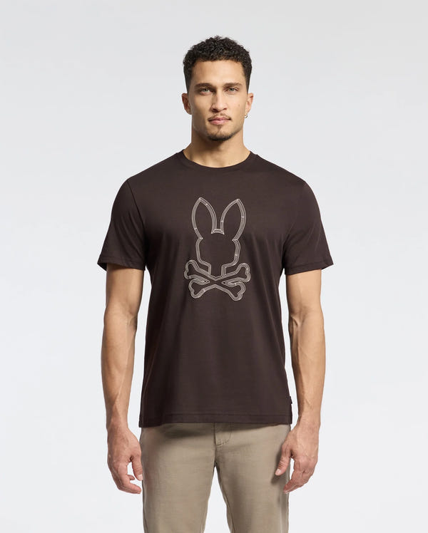 A man wearing a Psycho Bunny MENS MONARC FRONT BUNNY GRAPHIC TEE - B6U211E200, made from Pima cotton, stands against a light gray backdrop. The black tee showcases a bunny with crossbones design. With short curly hair and beige pants, he completes his casual yet distinctive look.