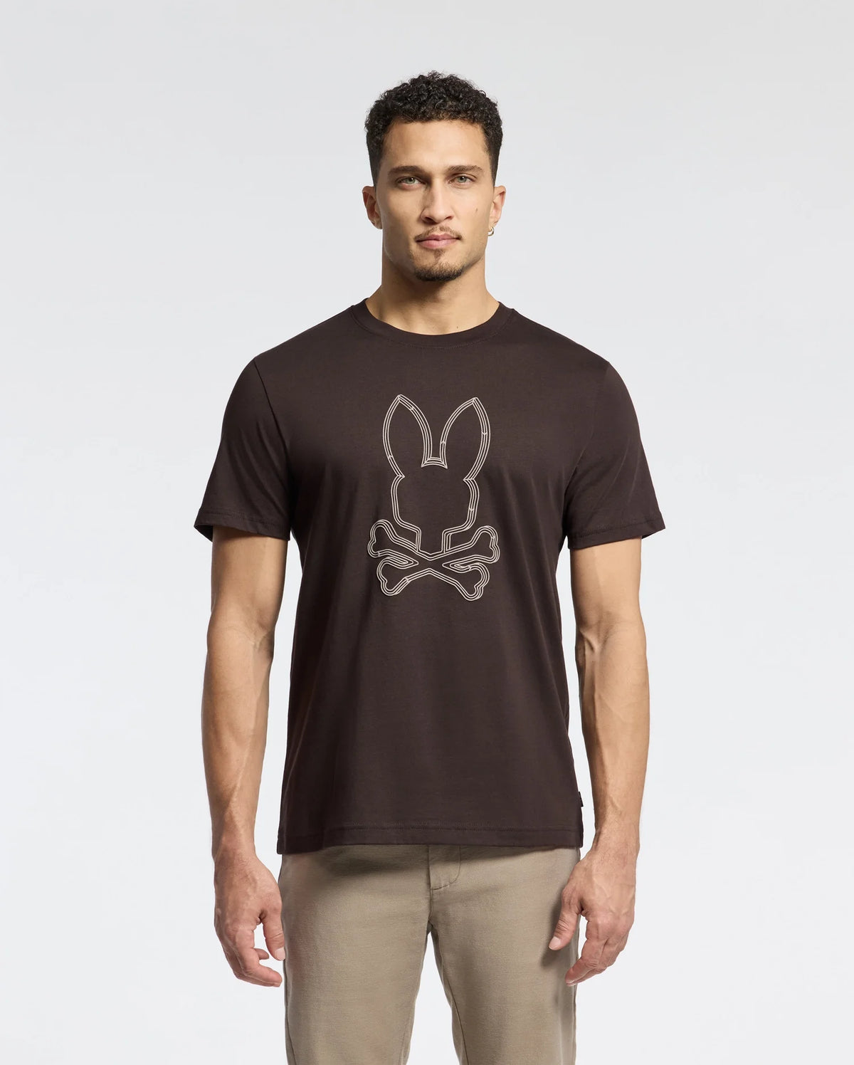 A man wearing a Psycho Bunny MENS MONARC FRONT BUNNY GRAPHIC TEE - B6U211E200, made from Pima cotton, stands against a light gray backdrop. The black tee showcases a bunny with crossbones design. With short curly hair and beige pants, he completes his casual yet distinctive look.