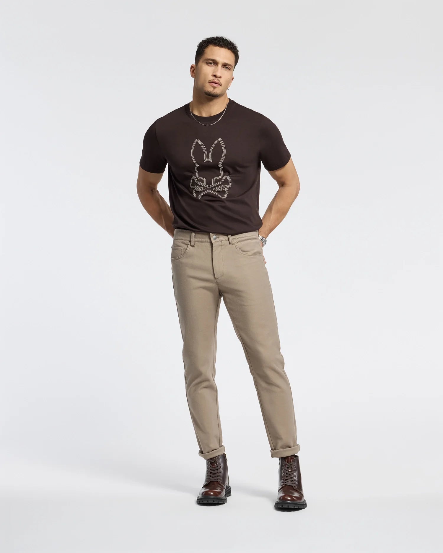 A person stands against a plain background wearing a dark Psycho Bunny B6U211E200 tee made from 100% Pima cotton, light-colored pants, and dark boots.