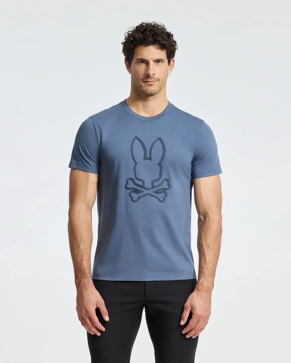 A man with curly hair wears a Psycho Bunny MENS MONARC FRONT BUNNY GRAPHIC TEE made from 100% Pima cotton. He pairs it with black pants and stands stylishly against a white background.
