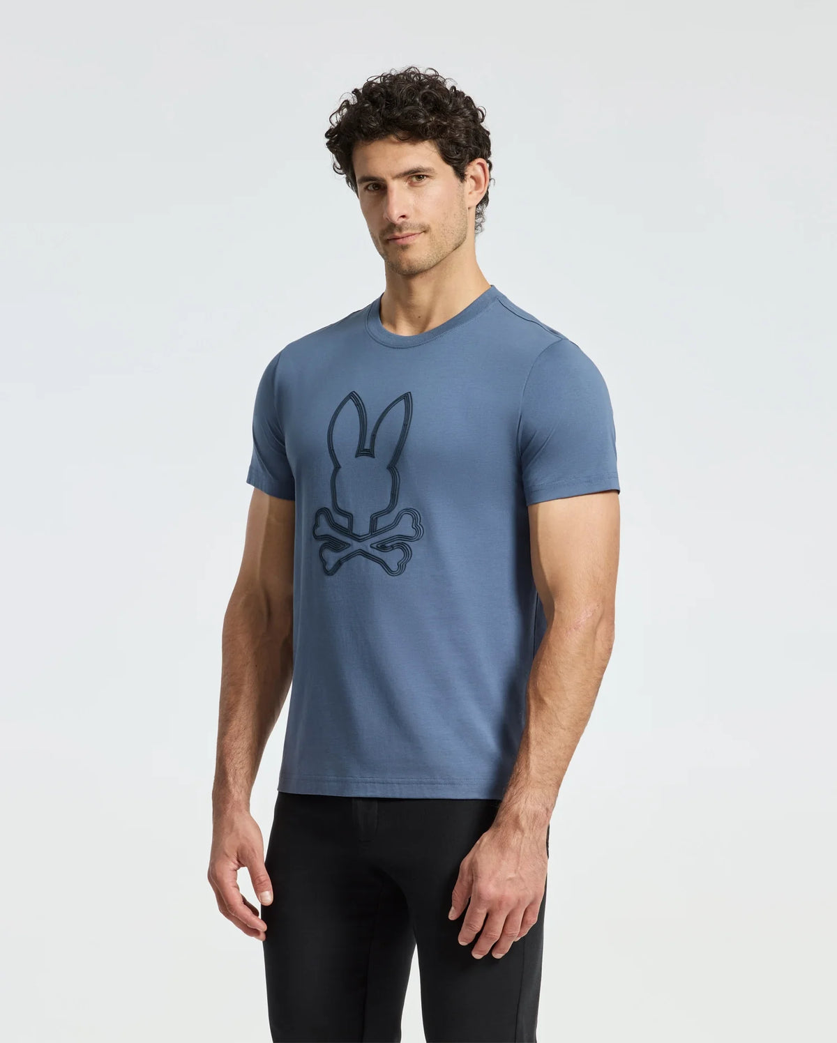 A man poses against a white background, wearing black pants and the Psycho Bunny MENS MONARC FRONT BUNNY GRAPHIC TEE - B6U211E200. Made from 100% Pima cotton, this blue tee features a stylish bunny and crossbones design, ensuring comfort and durability for any casual outing.