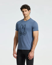 A man poses against a white background, wearing black pants and the Psycho Bunny MENS MONARC FRONT BUNNY GRAPHIC TEE - B6U211E200. Made from 100% Pima cotton, this blue tee features a stylish bunny and crossbones design, ensuring comfort and durability for any casual outing.
