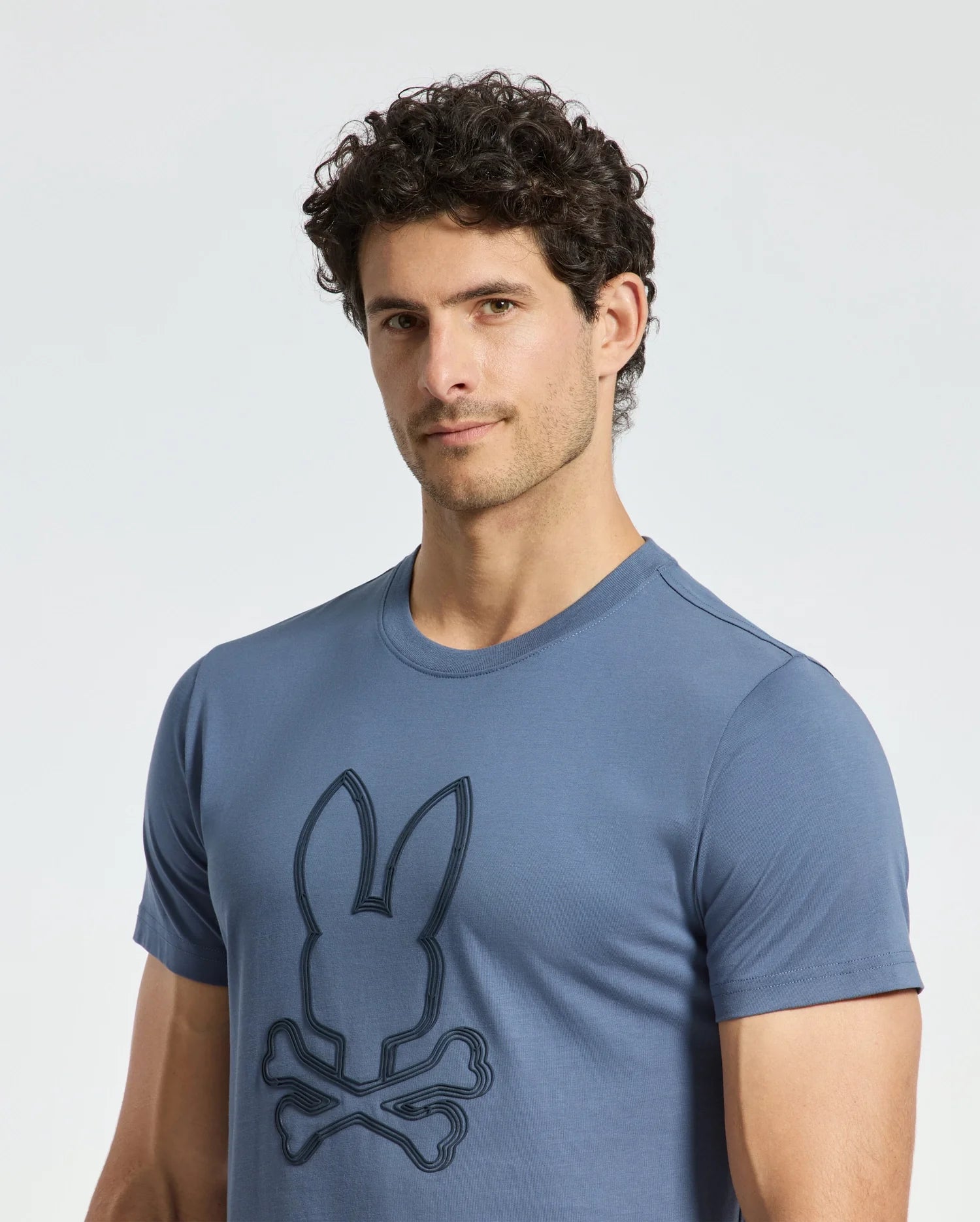 A man with curly hair confidently wears a Psycho Bunny MENS MONARC FRONT BUNNY GRAPHIC TEE - B6U211E200, made of 100% Pima cotton and showcasing a rabbit and crossbones design. He stands against a plain background, looking at the camera.
