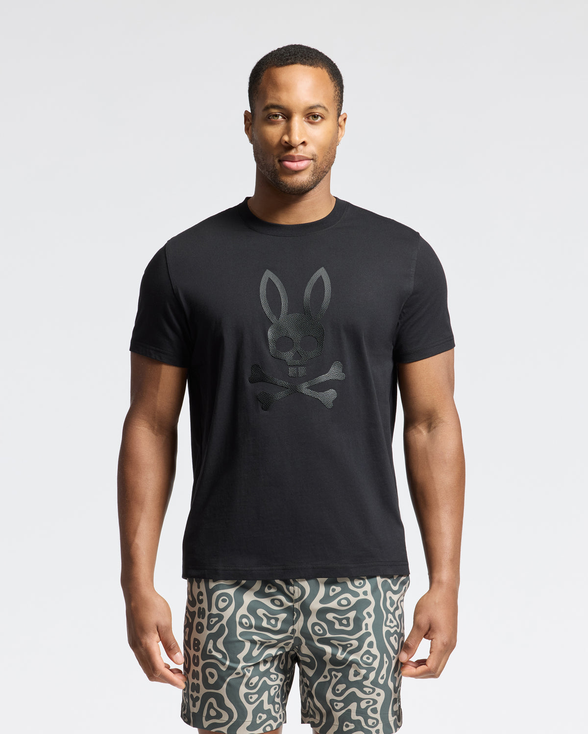 A man stands against a plain background wearing Psycho Bunny's black graphic Tee, MENS PAYSON LEATHER PRINT - B6U213E200, with a rabbit skull and crossbones. The Pima cotton jersey pairs effortlessly with his wavy patterned shorts for an effortlessly stylish look.