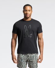 A man stands against a plain background wearing Psycho Bunny's black graphic Tee, MENS PAYSON LEATHER PRINT - B6U213E200, with a rabbit skull and crossbones. The Pima cotton jersey pairs effortlessly with his wavy patterned shorts for an effortlessly stylish look.