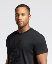 A man with short hair is wearing the Psycho Bunny MENS PAYSON LEATHER PRINT GRAPHIC TEE (B6U213E200), featuring a stylized rabbit skull and crossbones design. He stands against a plain background, gazing slightly to the side.