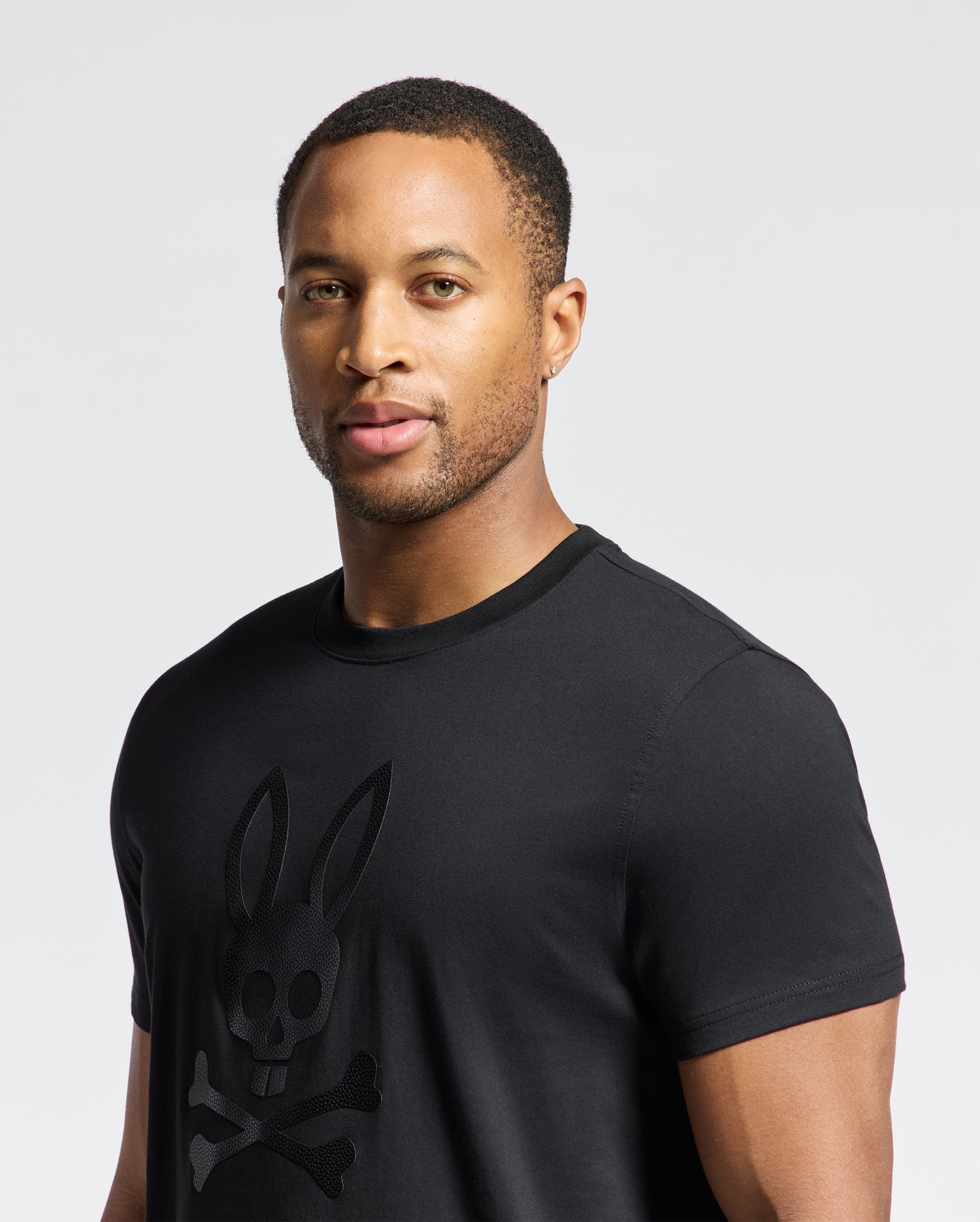 A man with short hair is wearing the Psycho Bunny MENS PAYSON LEATHER PRINT GRAPHIC TEE (B6U213E200), featuring a stylized rabbit skull and crossbones design. He stands against a plain background, gazing slightly to the side.