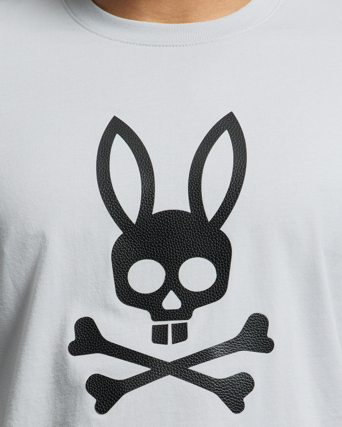 A close-up of the Psycho Bunny MENS PAYSON LEATHER PRINT GRAPHIC TEE - B6U213E200 features a gray Pima cotton jersey shirt with a black skull with rabbit ears, and two crossed bones in a playful pirate design highlighted by detailed leather print.