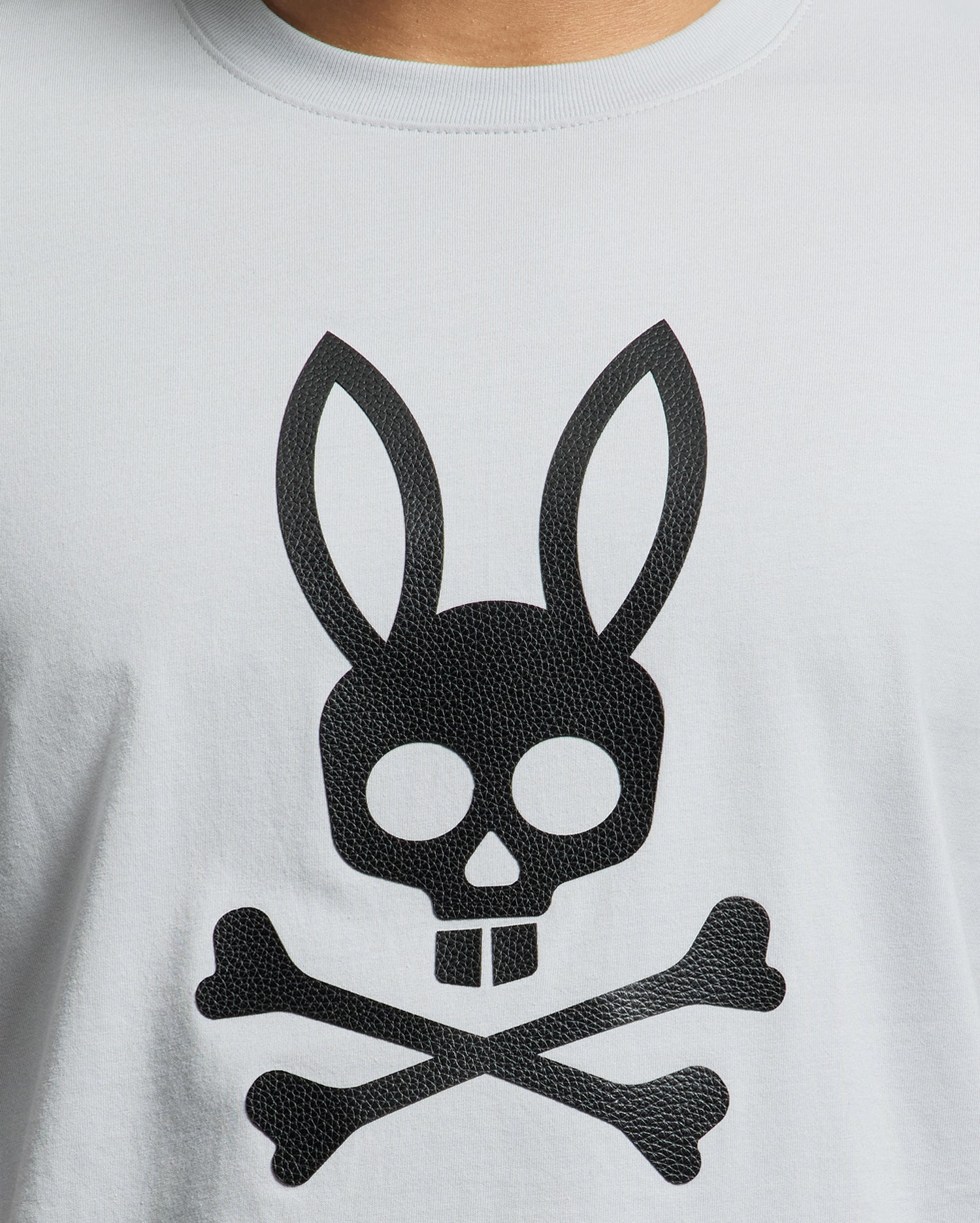 A close-up of the Psycho Bunny MENS PAYSON LEATHER PRINT GRAPHIC TEE - B6U213E200 features a gray Pima cotton jersey shirt with a black skull with rabbit ears, and two crossed bones in a playful pirate design highlighted by detailed leather print.