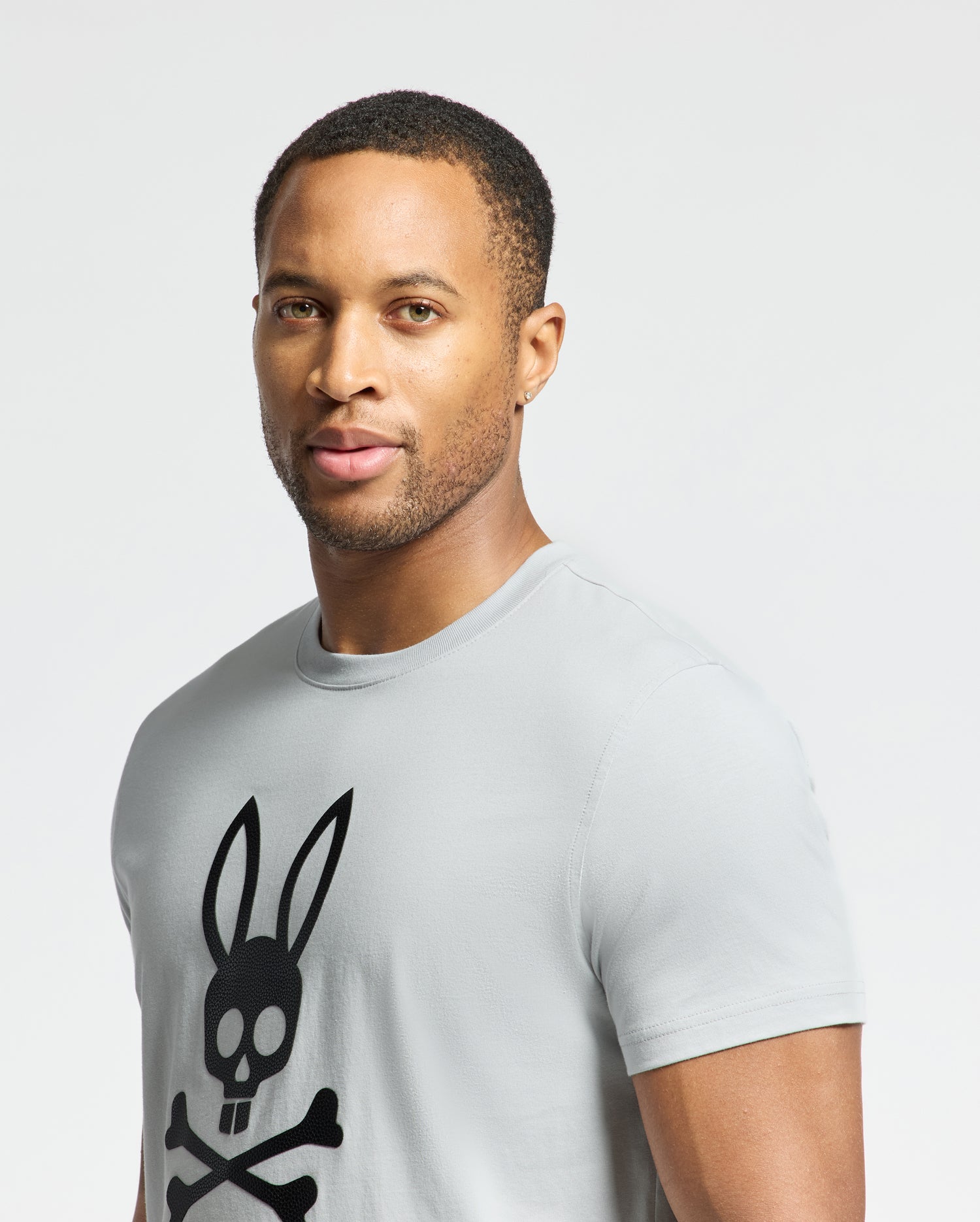 A man stands against a white background wearing the MENS PAYSON LEATHER PRINT GRAPHIC TEE - B6U213E200 by Psycho Bunny, featuring a black skull with rabbit ears design on light gray Pima cotton jersey.