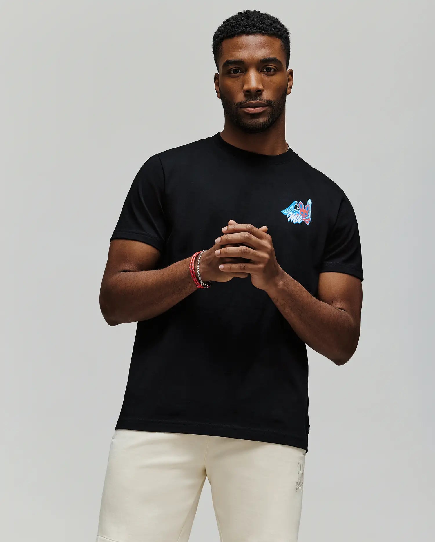 A man stands against a plain background wearing the Psycho Bunny MENS MONTREAL TEE (B6U381F200), a soft black Pima cotton t-shirt with a small colorful chest design, paired with light shorts. He clasps his hands and looks at the camera.