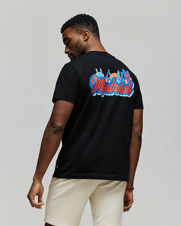 A man in the Psycho Bunny MENS MONTREAL TEE (B6U381F200), a black Pima cotton T-shirt with colorful Montreal-themed graphics, and white shorts stands facing away, turning his head to the side against a plain light gray background.