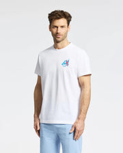 A man with short brown hair and a beard wears the Psycho Bunny MENS MONTREAL TEE - B6U381F200, featuring a small colorful mountain graphic on the left chest, and light blue pants, standing against a plain white background.