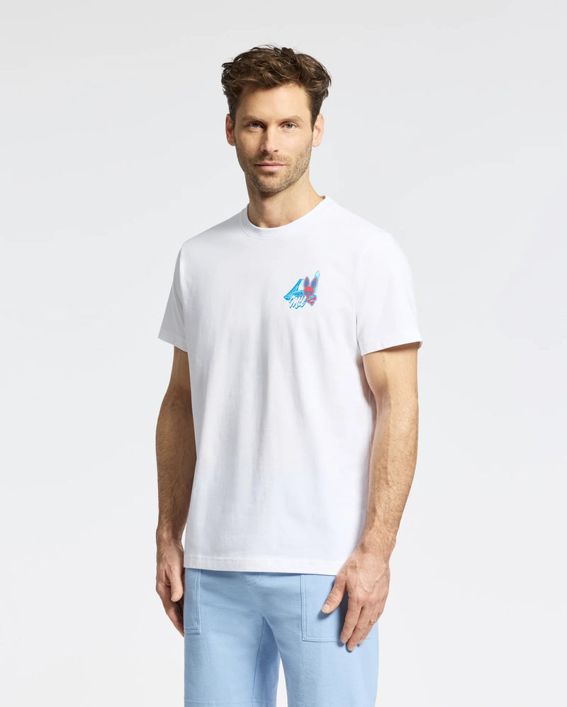 A man with short brown hair and a beard wears the Psycho Bunny MENS MONTREAL TEE - B6U381F200, featuring a small colorful mountain graphic on the left chest, and light blue pants, standing against a plain white background.