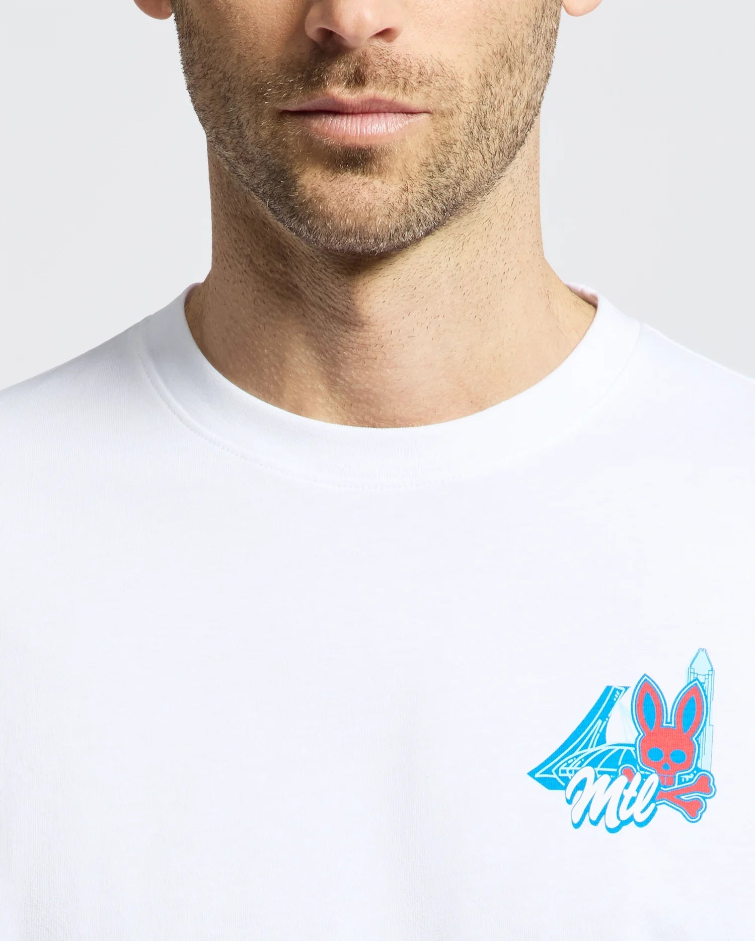 A man’s lower face and upper torso are visible as he wears the Psycho Bunny MENS MONTREAL TEE (B6U381F200), a white Pima cotton shirt with a small blue and pink chest graphic showing the number 4, bunny face, and “Mil.”.