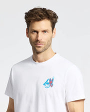 A man with short, messy brown hair and light stubble wears the Psycho Bunny MENS MONTREAL TEE (B6U381F200), a white Pima cotton shirt with a small, colorful cartoon graphic on the chest. He stands against a plain light background.