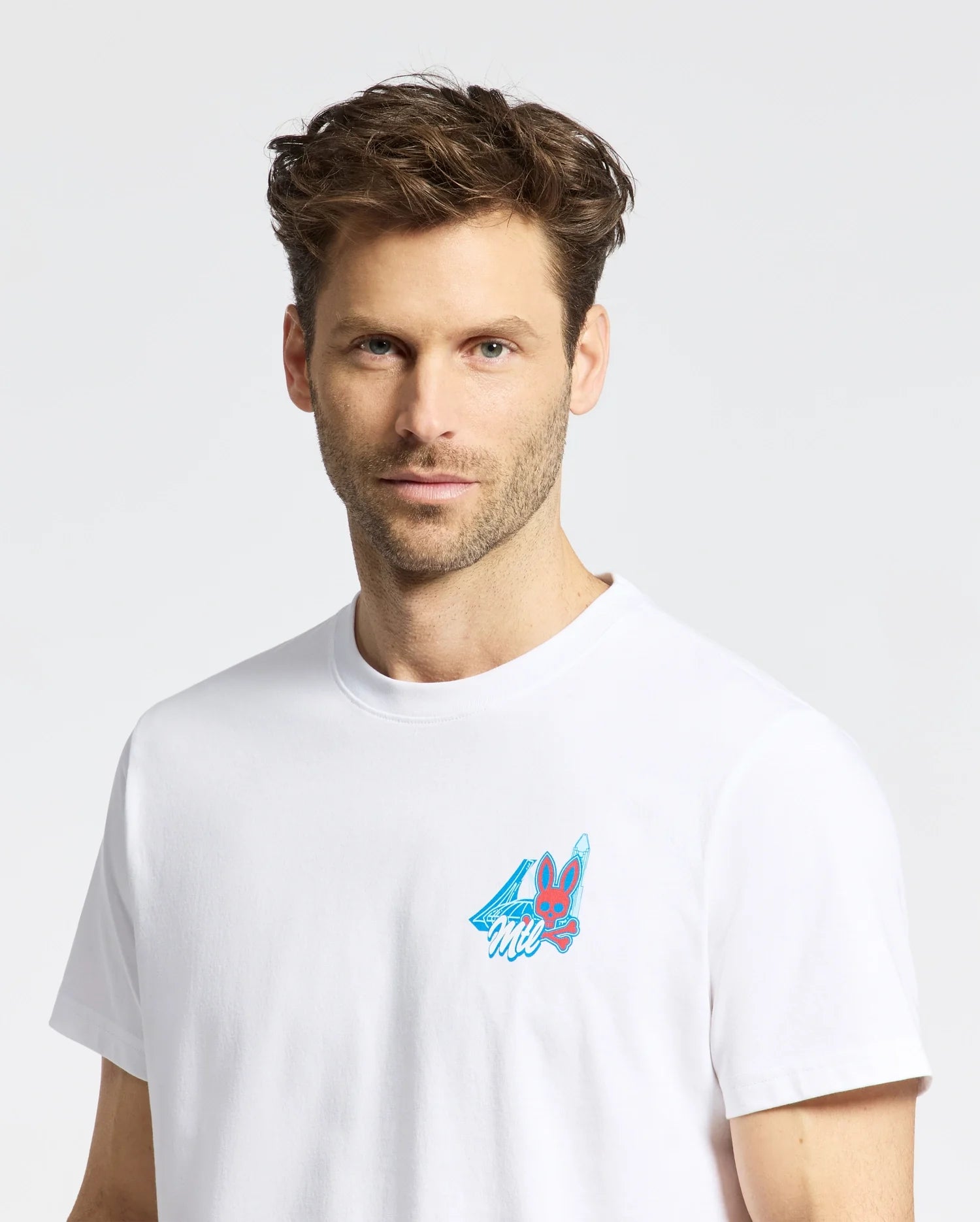 A man with short, messy brown hair and light stubble wears the Psycho Bunny MENS MONTREAL TEE (B6U381F200), a white Pima cotton shirt with a small, colorful cartoon graphic on the chest. He stands against a plain light background.