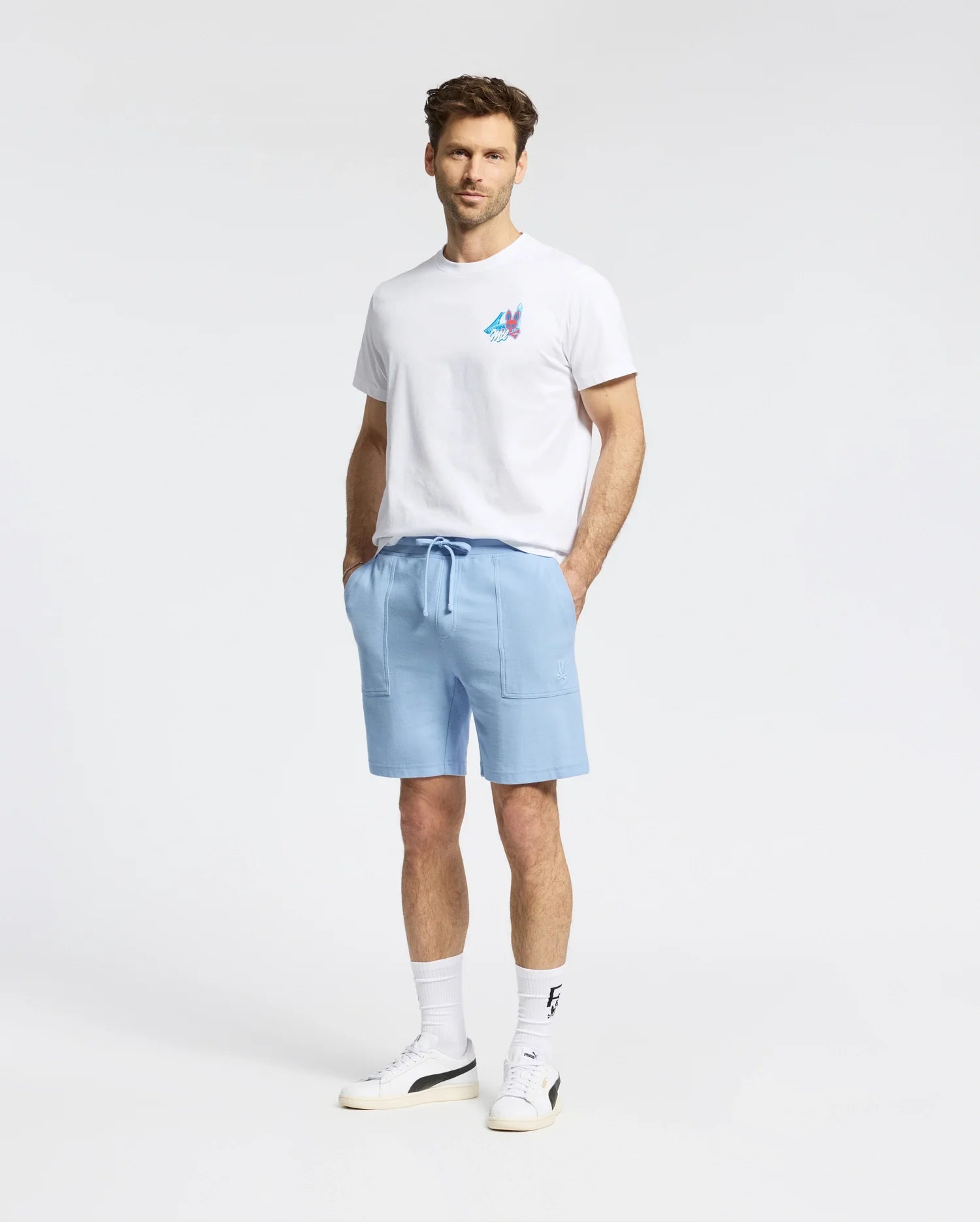 A man faces forward against a plain white background wearing a Psycho Bunny MENS MONTREAL TEE - B6U381F200 in white, paired with light blue drawstring shorts, white socks, and white sneakers.