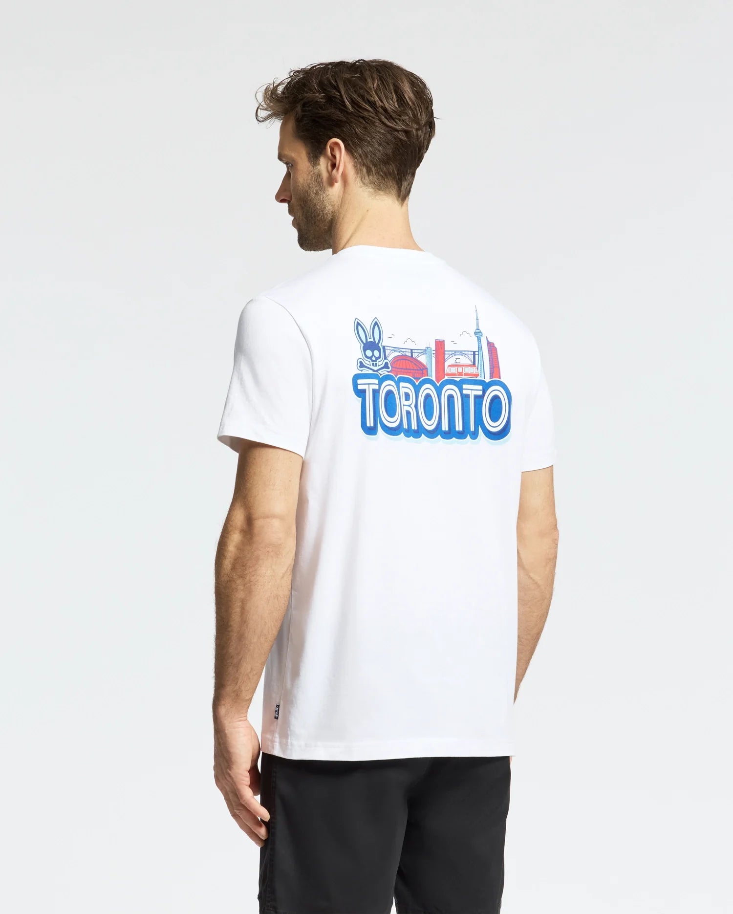 A man with short brown hair faces away, wearing Psycho Bunny’s MENS TORONTO TEE - B6U383F200 in soft Pima cotton, featuring colorful cartoon and city landmark graphics on the back. He pairs it with black shorts.