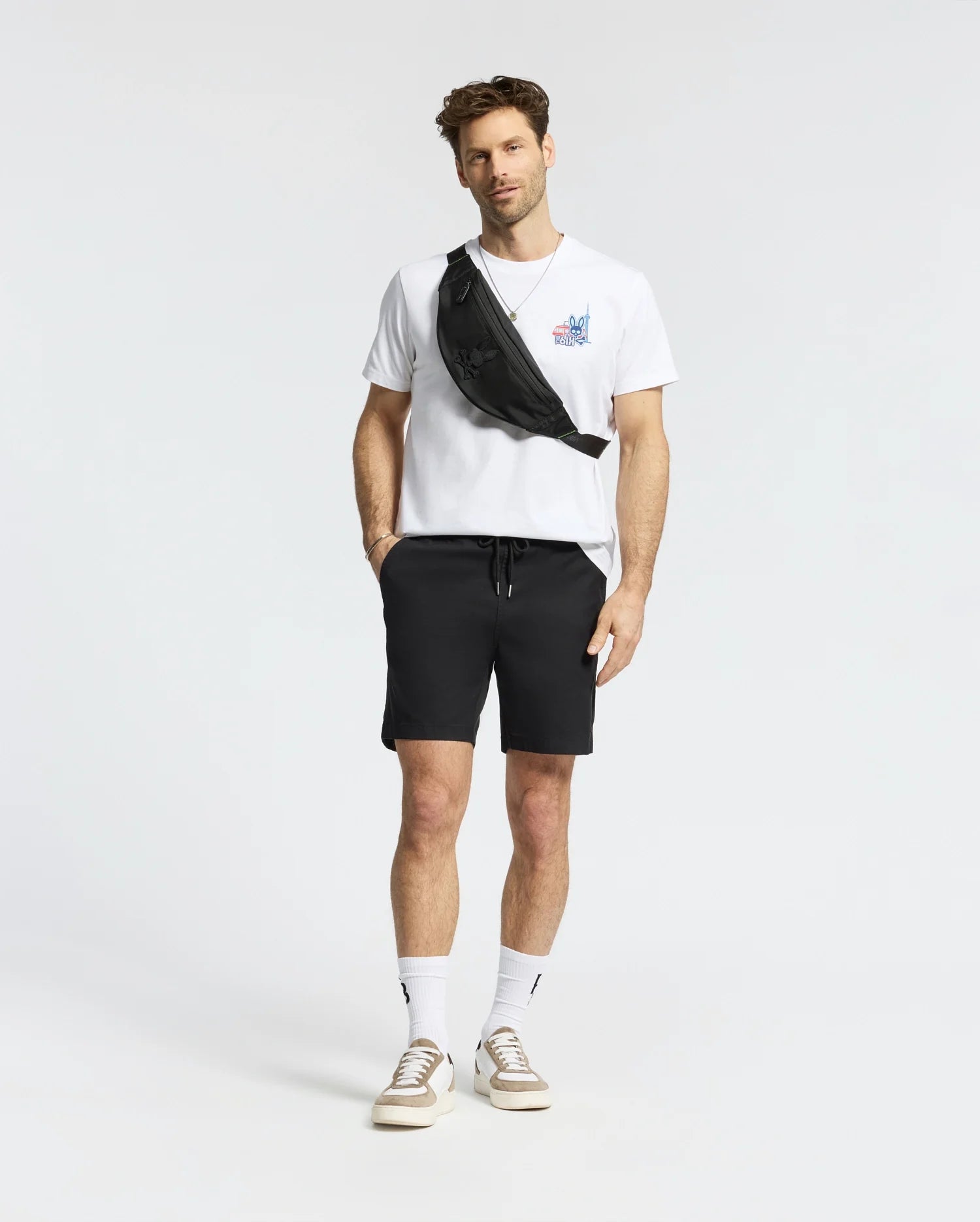 A man with short hair and a light beard stands against a white background, wearing the Psycho Bunny MENS TORONTO TEE - B6U383F200 in soft Pima cotton with bold graphics, black shorts, white socks, beige sneakers, and a black crossbody bag.
