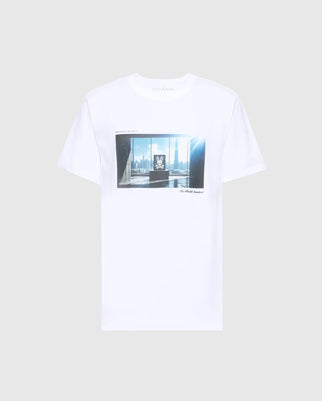 The MENS WARREN PHOTO REAL TEE by Psycho Bunny is a white Pima cotton T-shirt with a central graphic print of a city skyline through windows lit by sunlight, featuring illegible text.