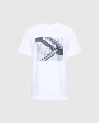 The MENS DEAN PHOTO REAL TEE - B6U390E200 by Psycho Bunny features a centered monochrome graphic of urban geometric shapes, a stairway, and plant leaves on a white Pima cotton jersey.