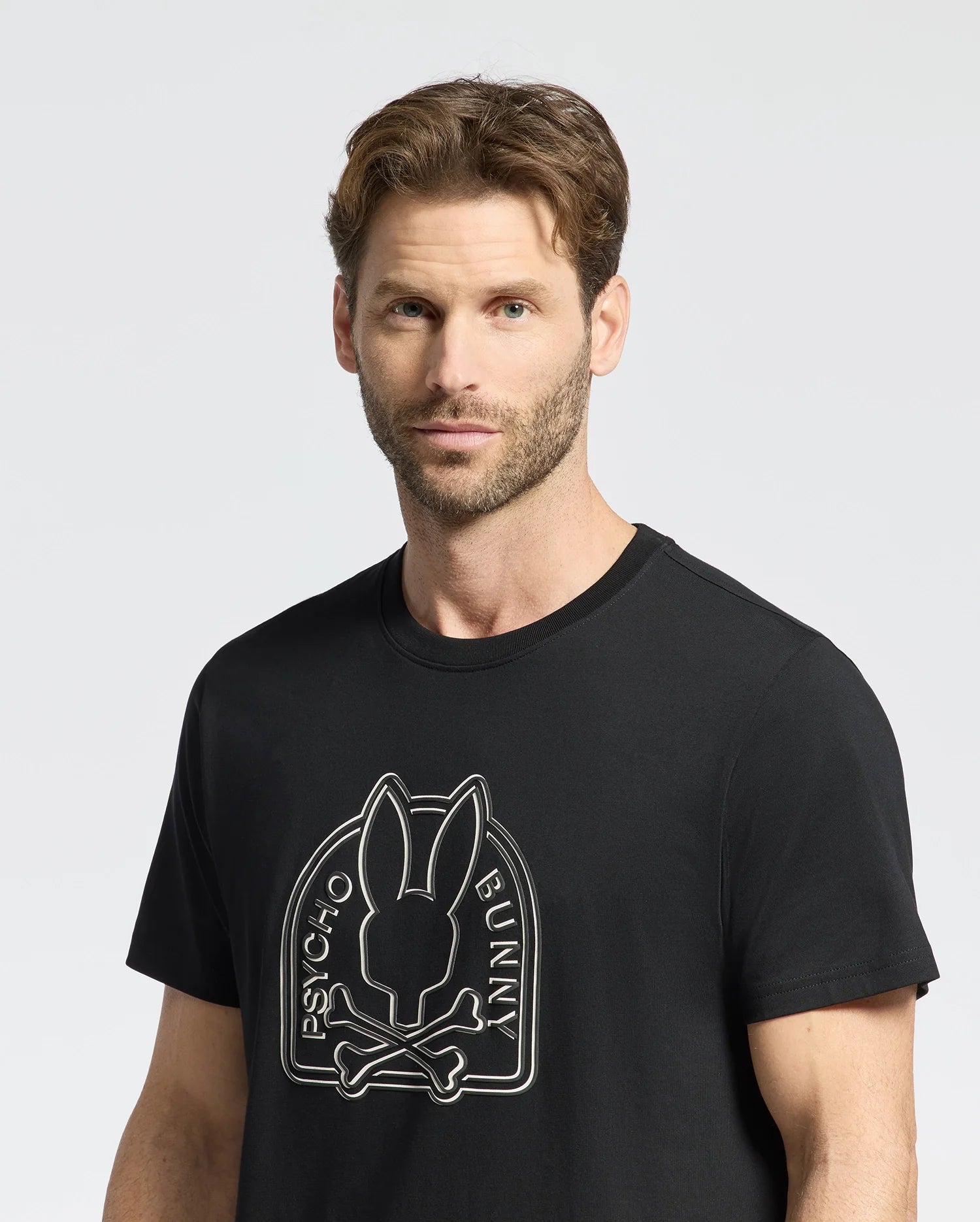 A man with short brown hair and a beard wears the edgy Psycho Bunny MENS MELBOURNE GRAPHIC TEE, B6U410F200. The black Pima Cotton shirt features a stylized bunny graphic with crossbones and "Psycho Bunny" text, standing out against a plain light gray background.