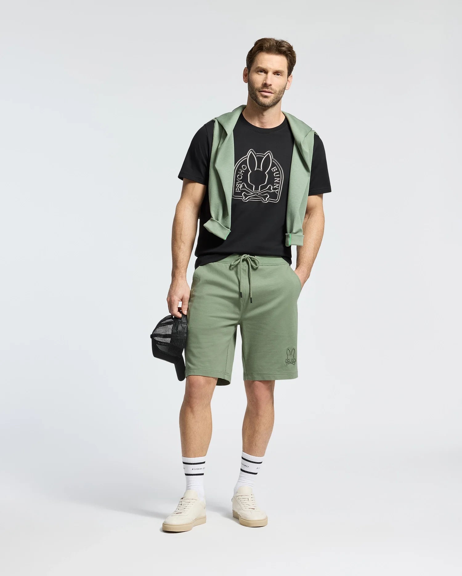 A man in a black Psycho Bunny MENS MELBOURNE GRAPHIC TEE, paired with green shorts and a green sweatshirt draped over his shoulders. He holds a black cap, completing the look with white sneakers and striped socks.
