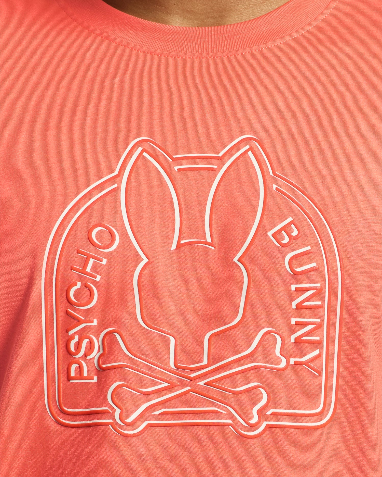 The Melbourne tee by Psycho Bunny, made from eco-friendly Pima Cotton in coral, features an embossed bunny head with crossbones design and "Psycho Bunny" printed on the sides.