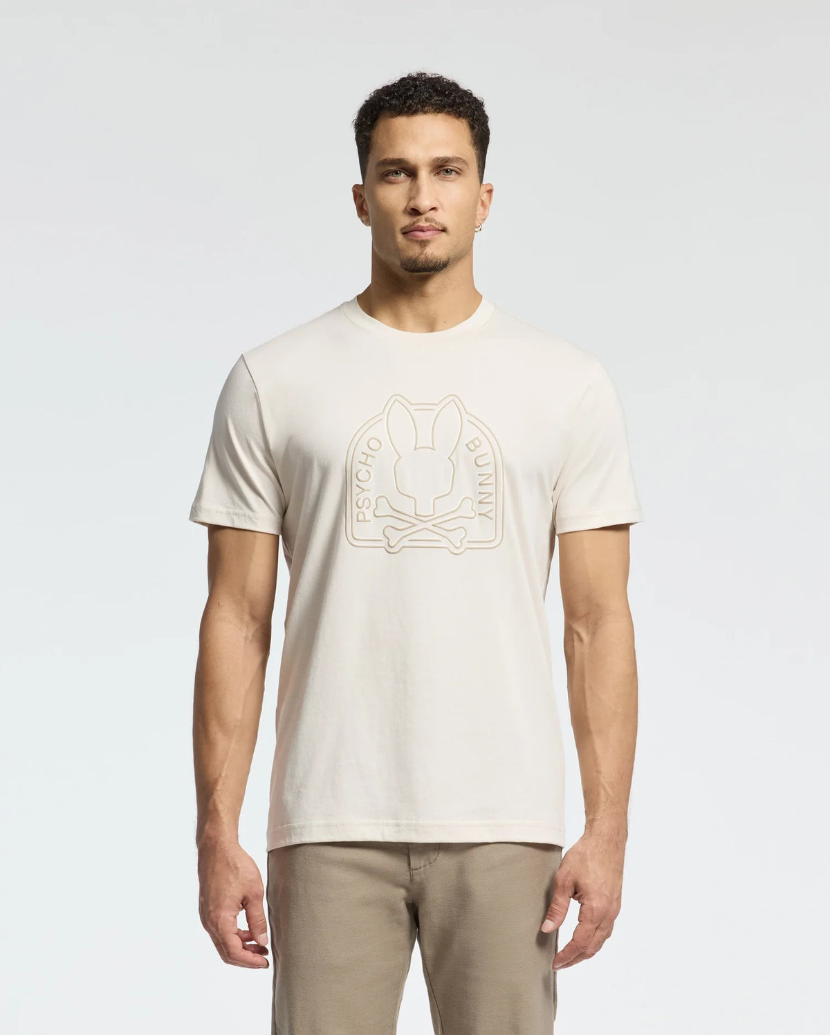 A man wears a cream MENS MELBOURNE GRAPHIC TEE - B6U410F200 by Psycho Bunny, showcasing a bunny logo and "Psycho Bunny" on the front. Made from eco-friendly Peruvian Pima cotton, he pairs it with beige pants against a plain white backdrop.