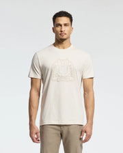 A man wears a cream MENS MELBOURNE GRAPHIC TEE - B6U410F200 by Psycho Bunny, showcasing a bunny logo and "Psycho Bunny" on the front. Made from eco-friendly Peruvian Pima cotton, he pairs it with beige pants against a plain white backdrop.