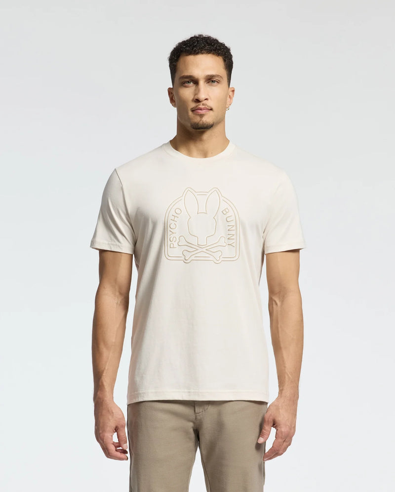 A man wears a cream MENS MELBOURNE GRAPHIC TEE - B6U410F200 by Psycho Bunny, showcasing a bunny logo and "Psycho Bunny" on the front. Made from eco-friendly Peruvian Pima cotton, he pairs it with beige pants against a plain white backdrop.